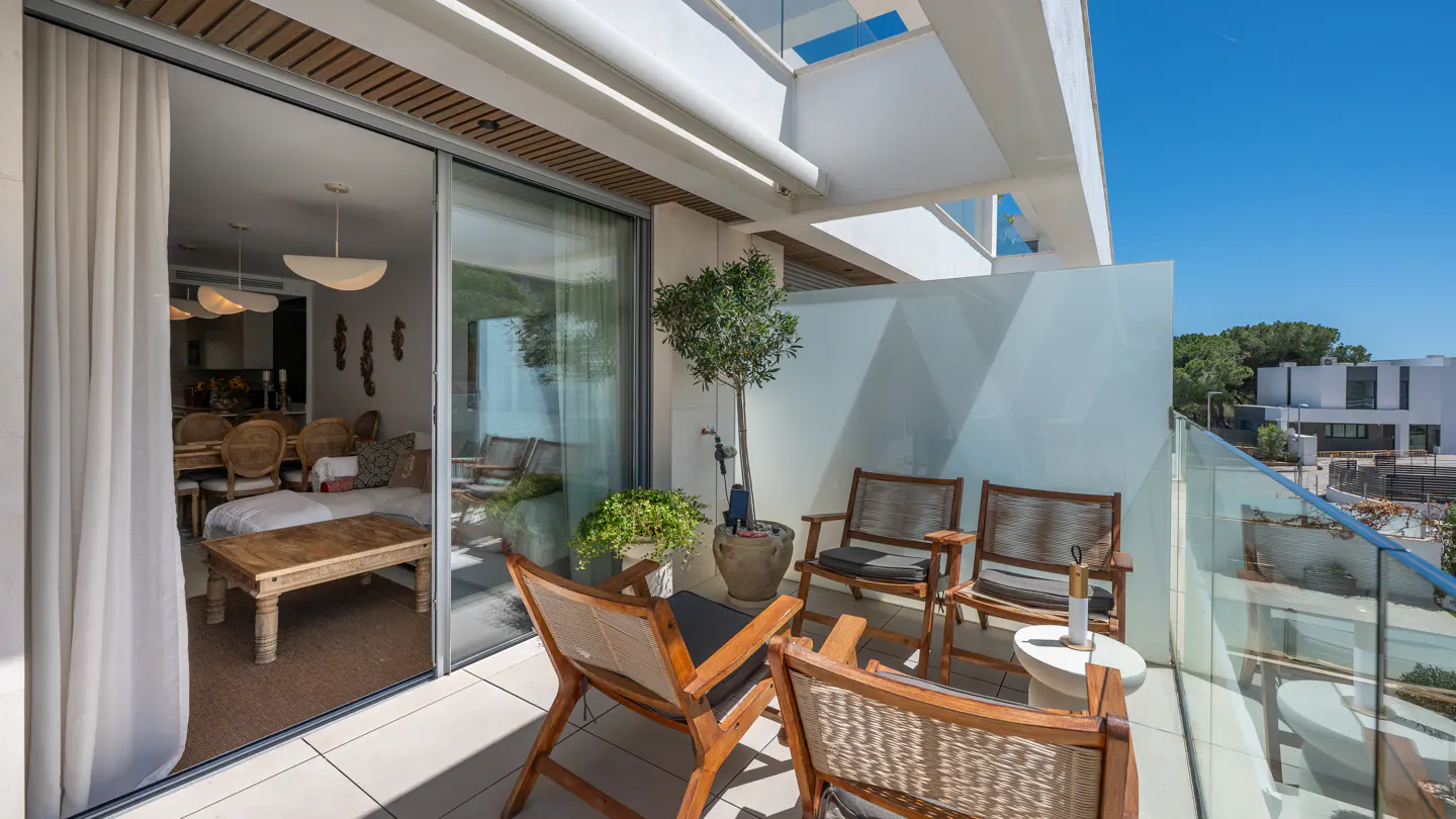 A bright balcony with wooden chairs, a small table, and potted plants. Sliding glass doors lead to an interior dining area.