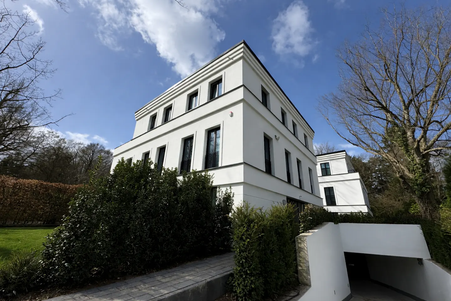 Exterior view of a modern, three-story white house with black-framed windows, surrounded by green bushes and trees under a blue sky.