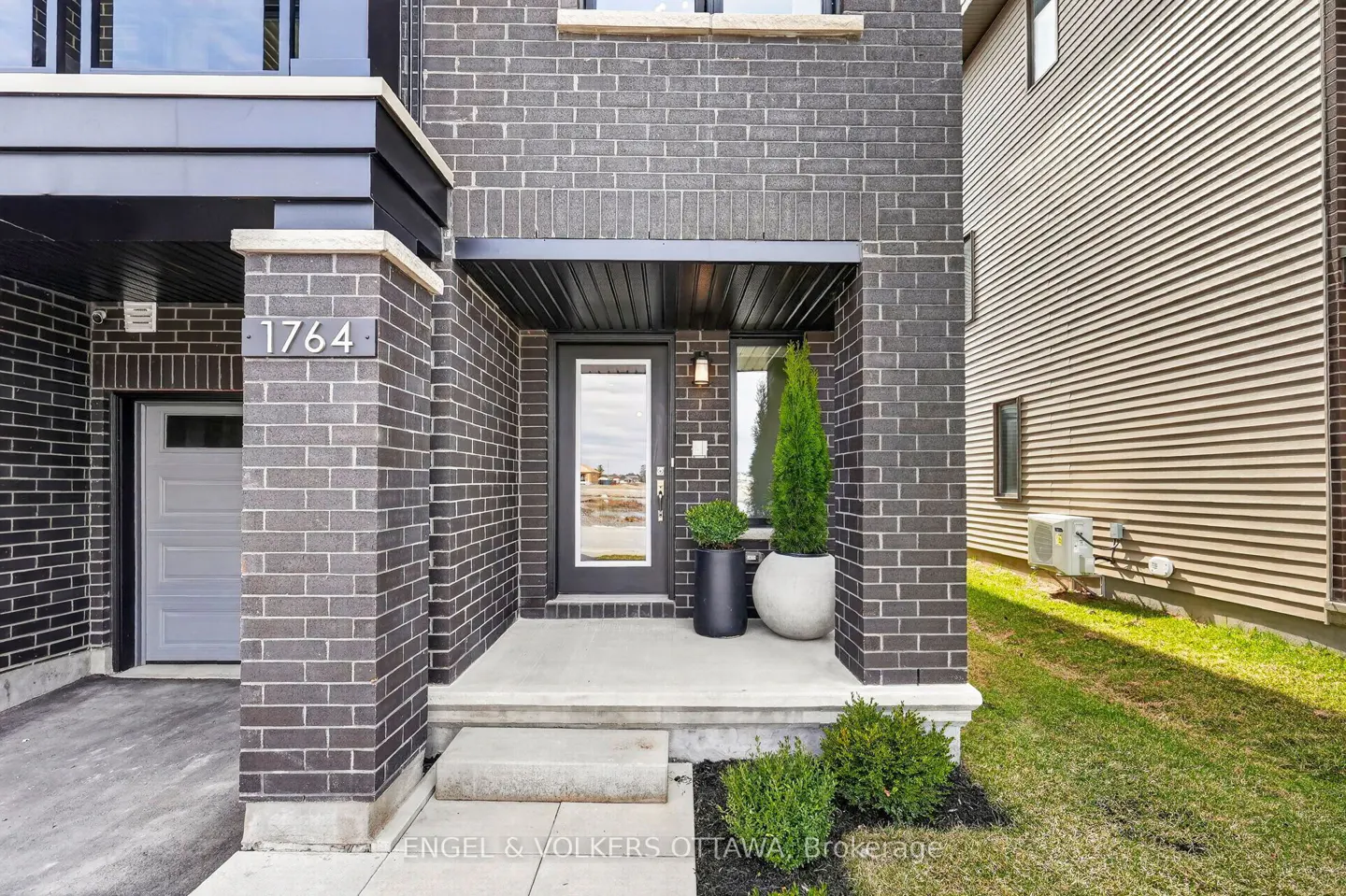Exterior of a modern brick home with the address 1764. A black-framed glass door is flanked by potted plants. A grey garage door is visible to the left.