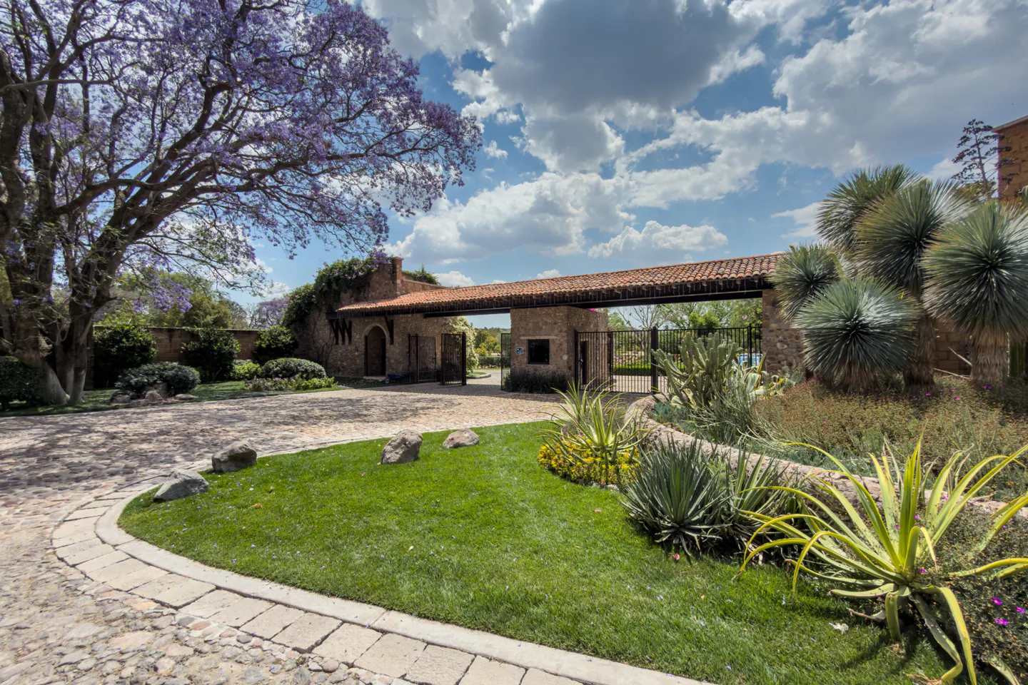 Gated entrance to a property with a red tile roof, stone walls, and lush landscaping with a flowering tree.