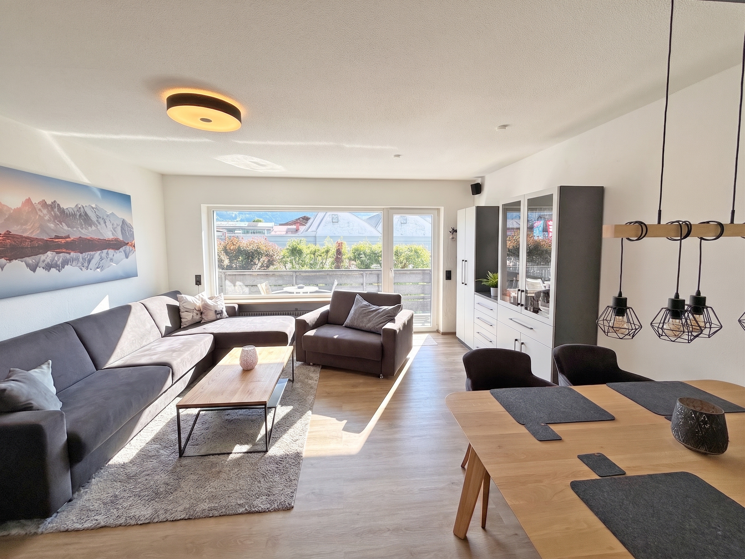 Bright open-plan living room with a grey sectional sofa, wooden coffee table, dining area, and large window with balcony. [AI-generated text] [search.image.alt.aiModified]