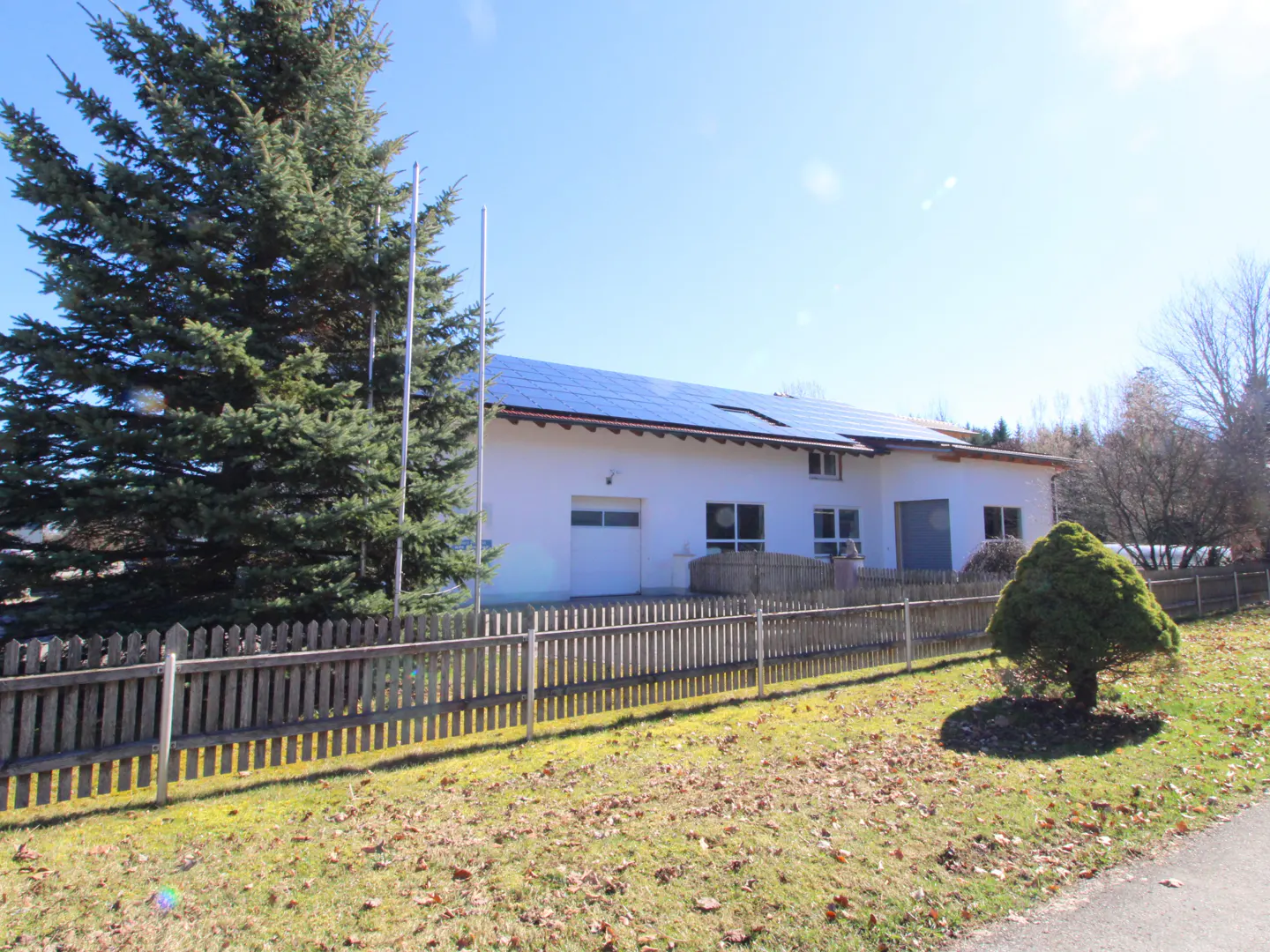 A white building with solar panels on the roof, a garage door, and a wooden fence in front. A large pine tree is on the left.