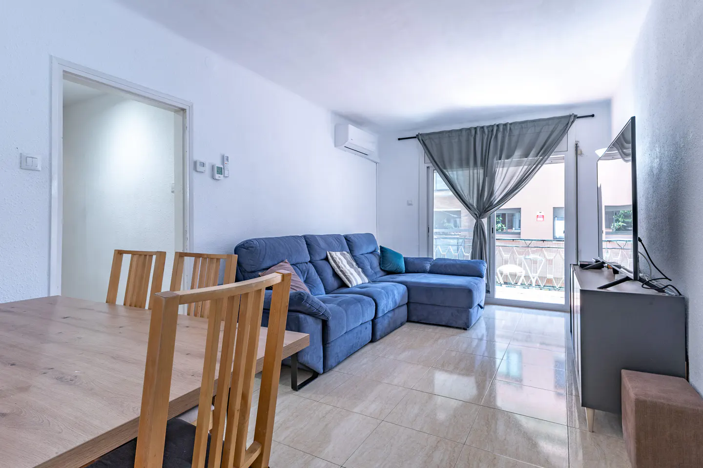 Bright living room with a blue sectional sofa, wood dining table, and a TV near a balcony with gray curtains.