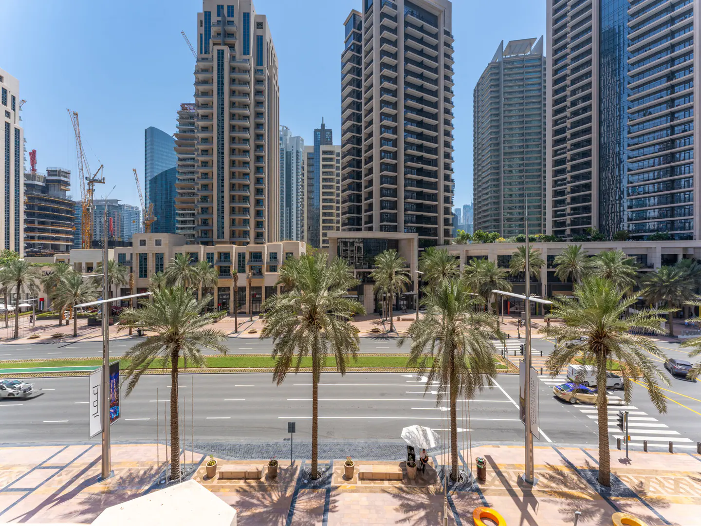 View of a city street lined with palm trees and tall buildings under a clear blue sky. Cars drive on the road.