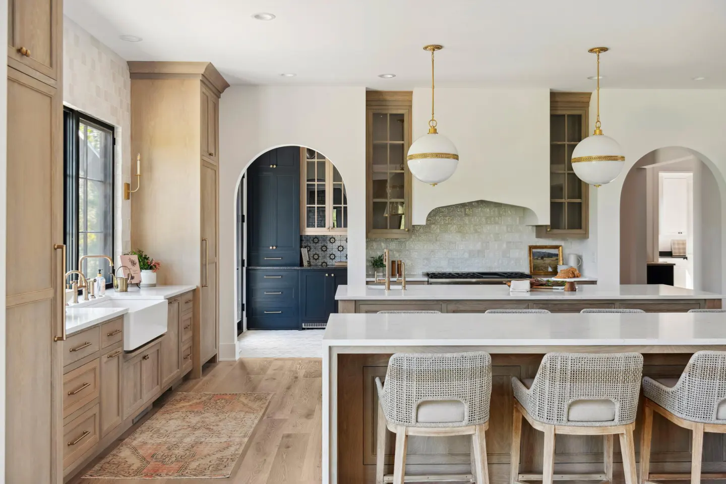 Bright, modern kitchen with wood floors, cabinets, and island with woven chairs. Globe pendant lights hang above the island. Arched doorways lead to other rooms.