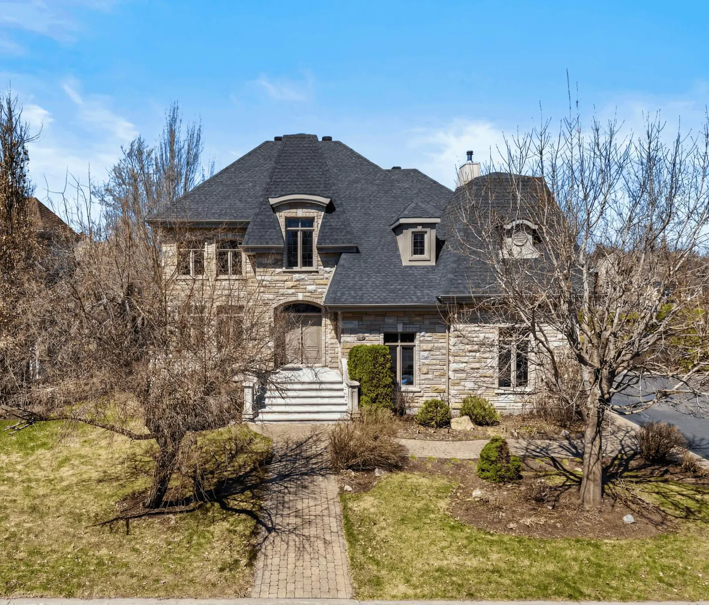 Two-story stone house with a dark roof, dormer windows, and a brick walkway leading to the front door.