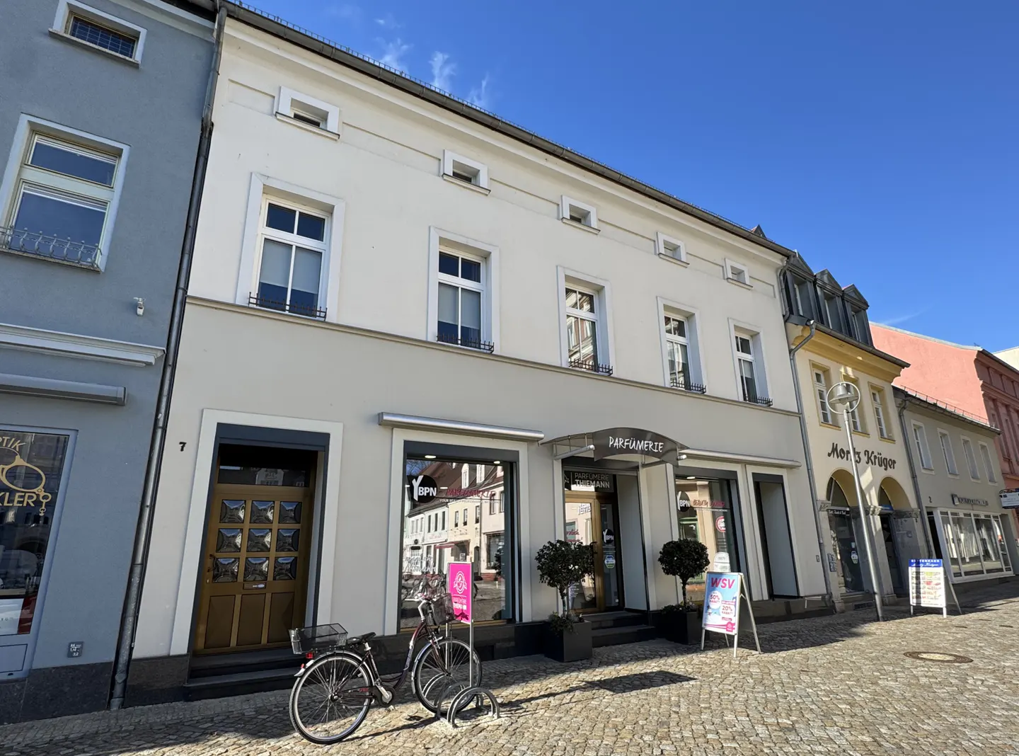 Street view of a three-story building with a "Parfumerie" shop on the ground floor. A bicycle is parked in front.