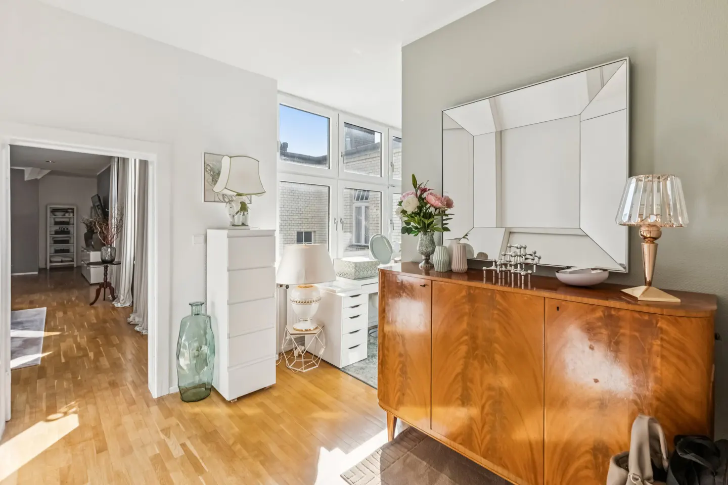 Hallway with wood floors, a brown cabinet with a mirror above, and a white dresser with a lamp. A doorway leads to another room.