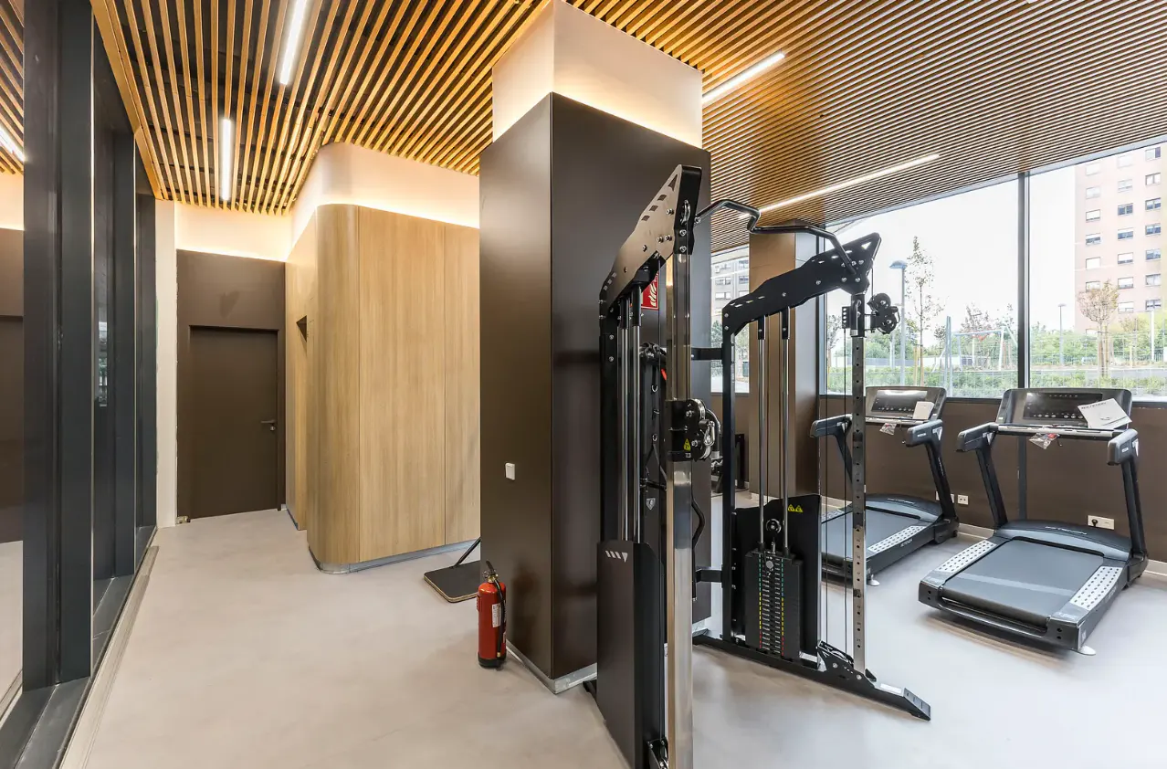 Modern apartment gym with treadmills, a cable machine, wood-slat ceiling, and large windows overlooking a city street.