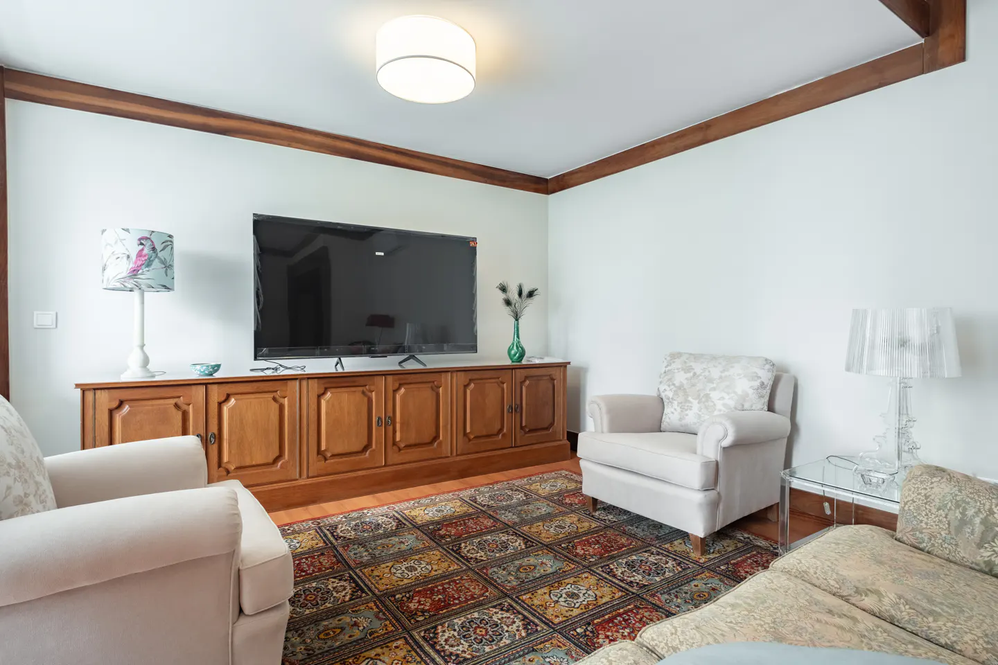 Living room with white walls, wood trim, and a patterned rug. A large TV sits atop a wooden cabinet. Two armchairs face the TV.