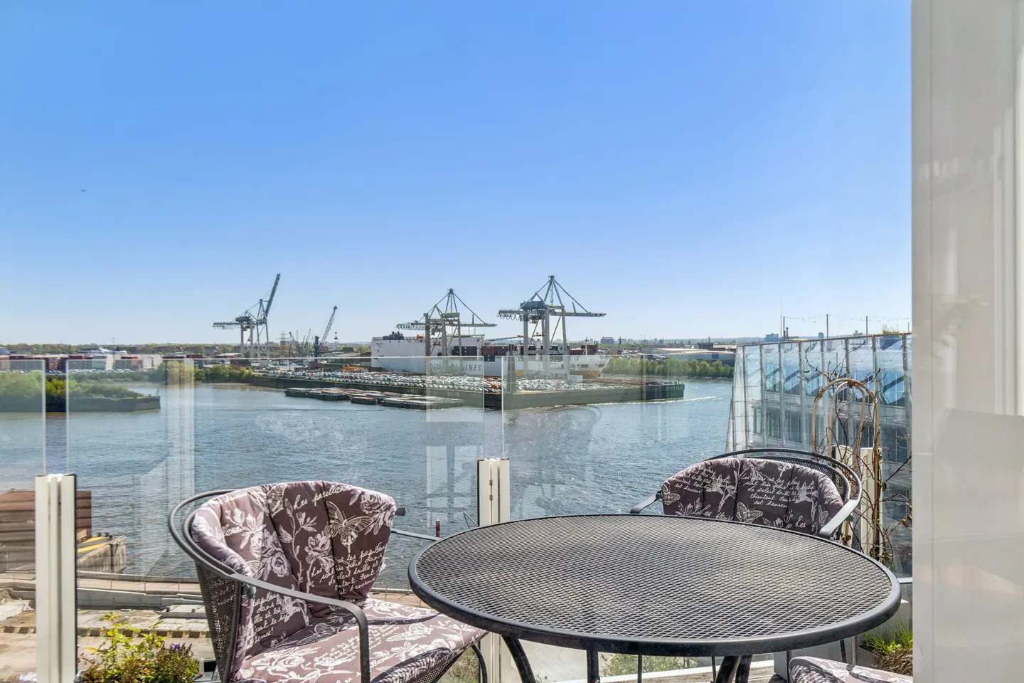 Balcony view with black table and chairs overlooking a river with cargo ships and cranes under a clear blue sky.