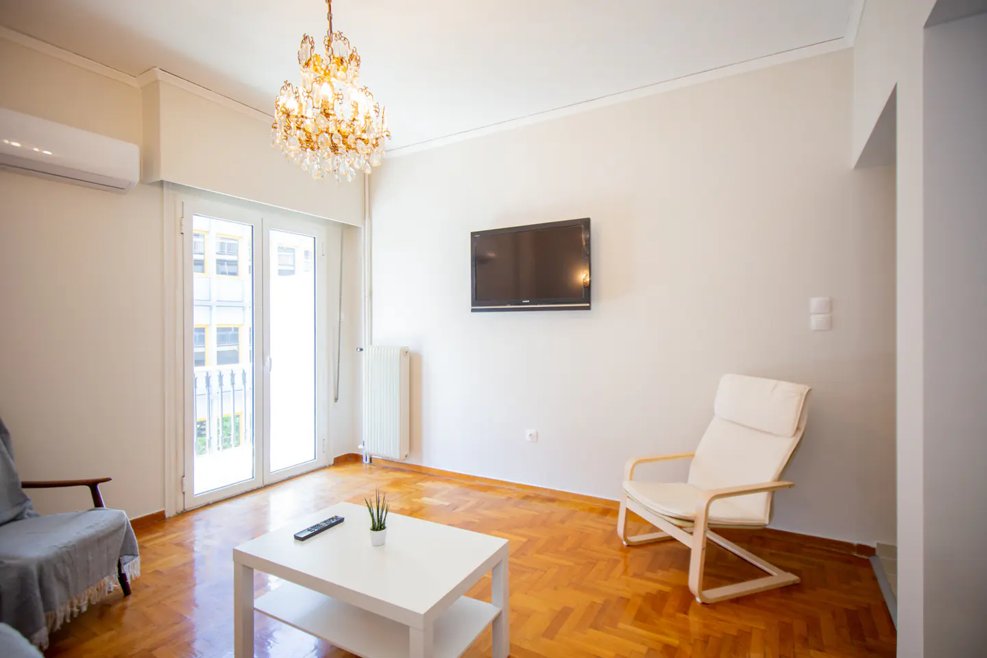Bright living room with wood floors, white walls, and a crystal chandelier. A TV hangs above a white chair. A white coffee table sits in front of a glass door.