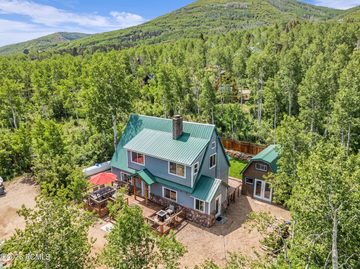 Aerial view of a blue house with a green roof, surrounded by trees and a mountain backdrop.