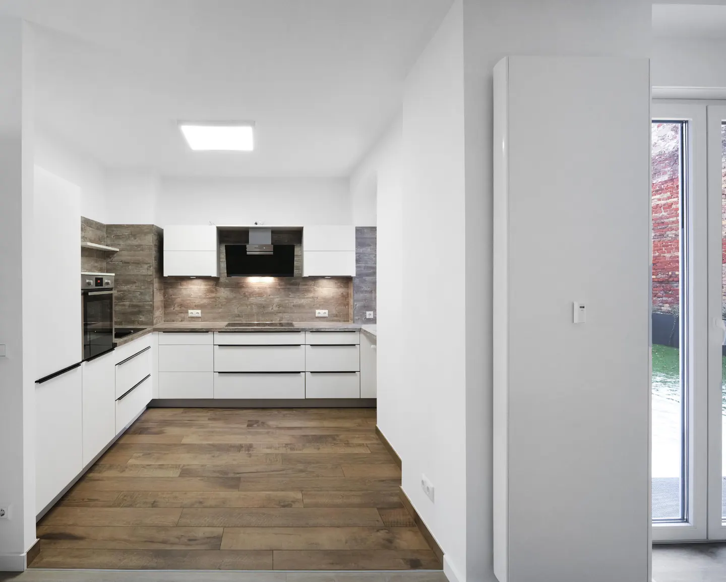 Bright, modern kitchen with white cabinets, wood floors, and stainless steel appliances. A window shows a brick wall and green lawn.