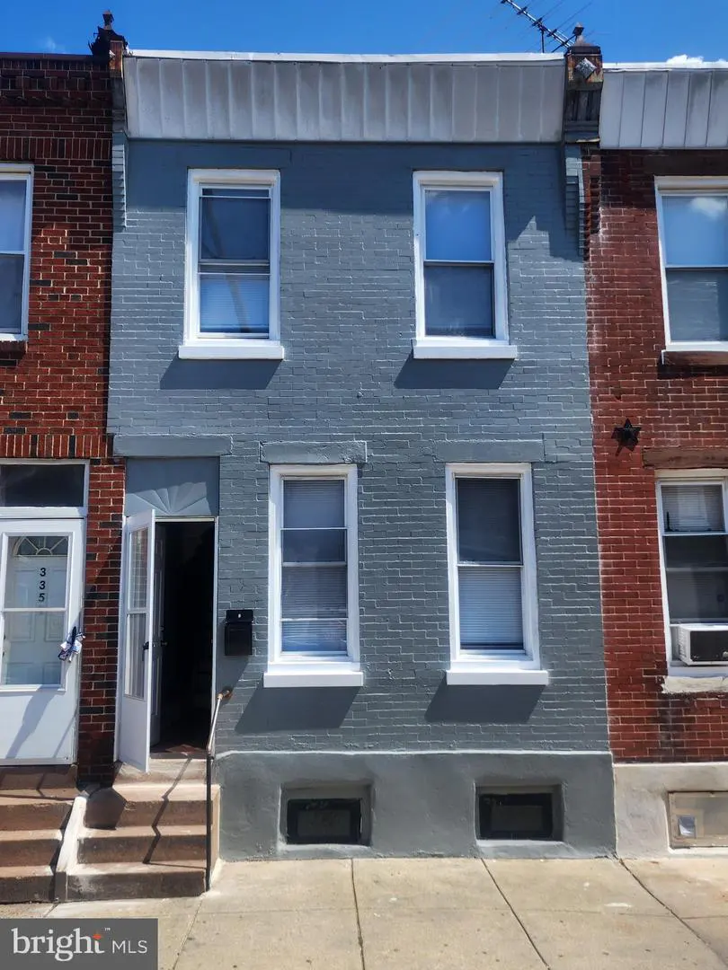 A two-story row house with a gray brick facade and white-framed windows under a blue sky. The front door is open.