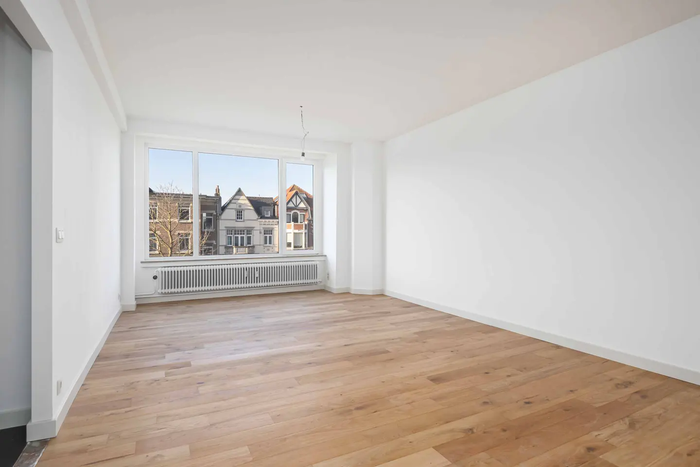 Bright, empty room with light wood floors and white walls. A large window shows houses across the street.