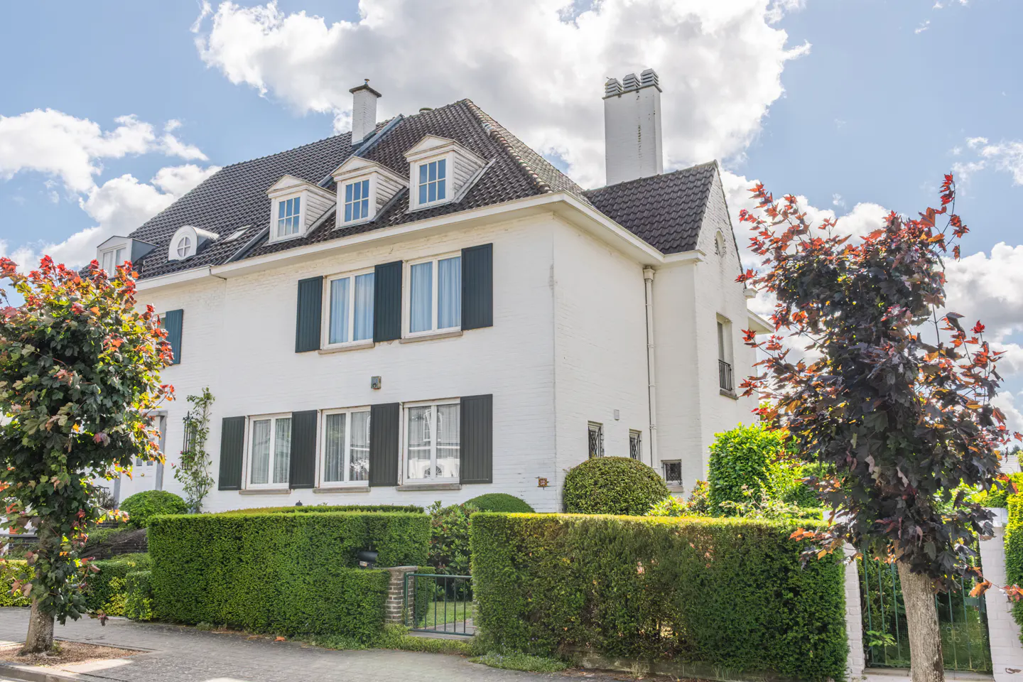 A white two-story house with dark shutters, a tiled roof, and dormer windows, surrounded by manicured hedges and trees.