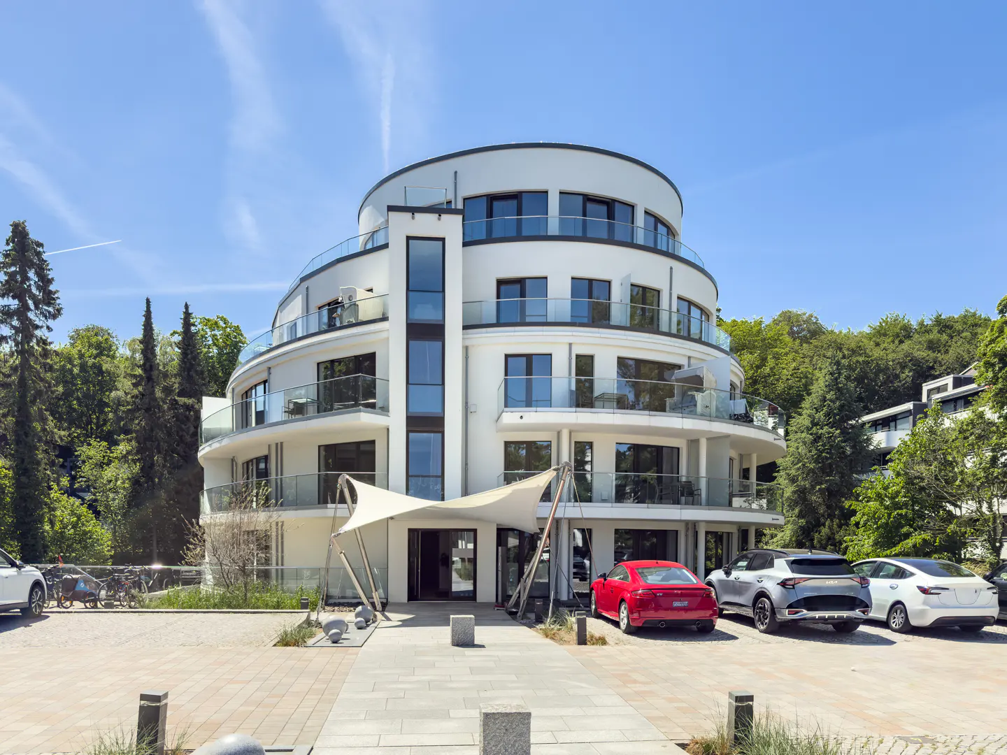 Modern white circular apartment building with glass balconies, a shaded entrance, and parked cars under a clear blue sky.