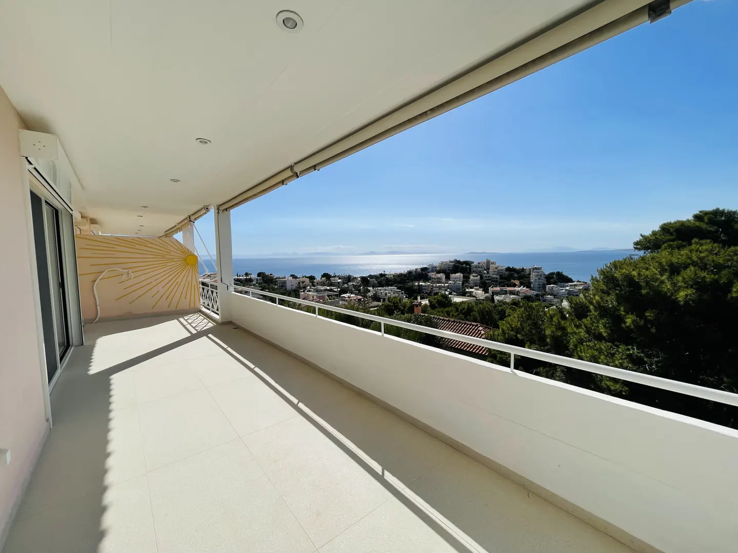 Wide balcony with white tile floor and railing overlooks a coastal town and the blue sea under a clear sky. A sun design is on the wall.