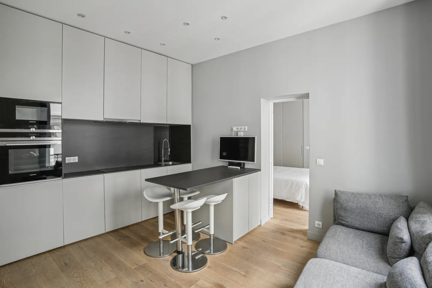 Modern apartment interior with gray cabinets, black countertops, and a breakfast bar with three white stools. A gray sofa is visible in the foreground.