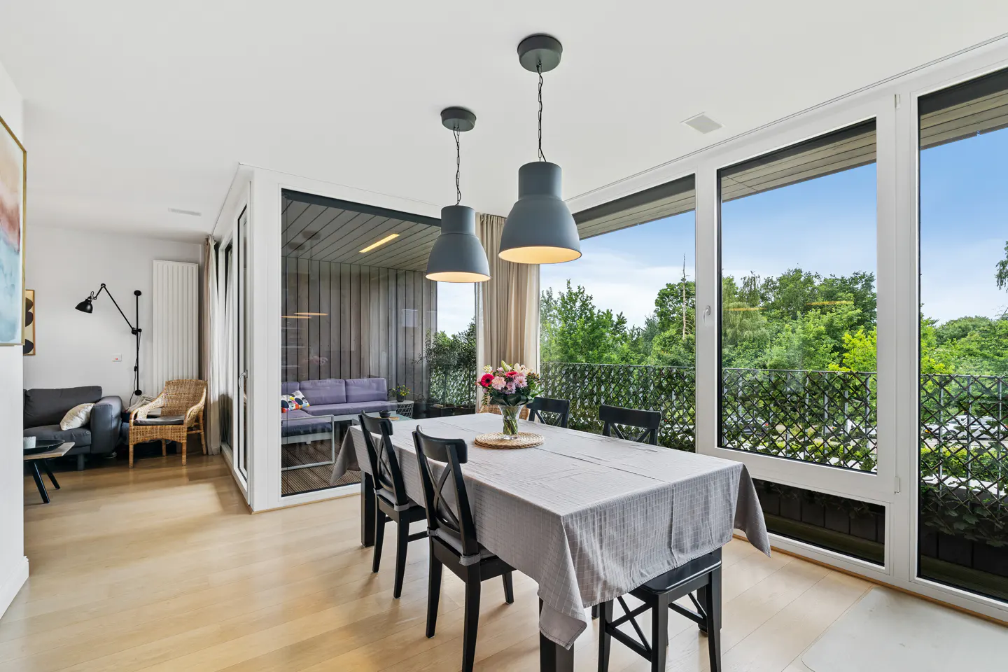 Dining area with a grey-clothed table, black chairs, and pendant lights, overlooking a balcony with lush green trees.