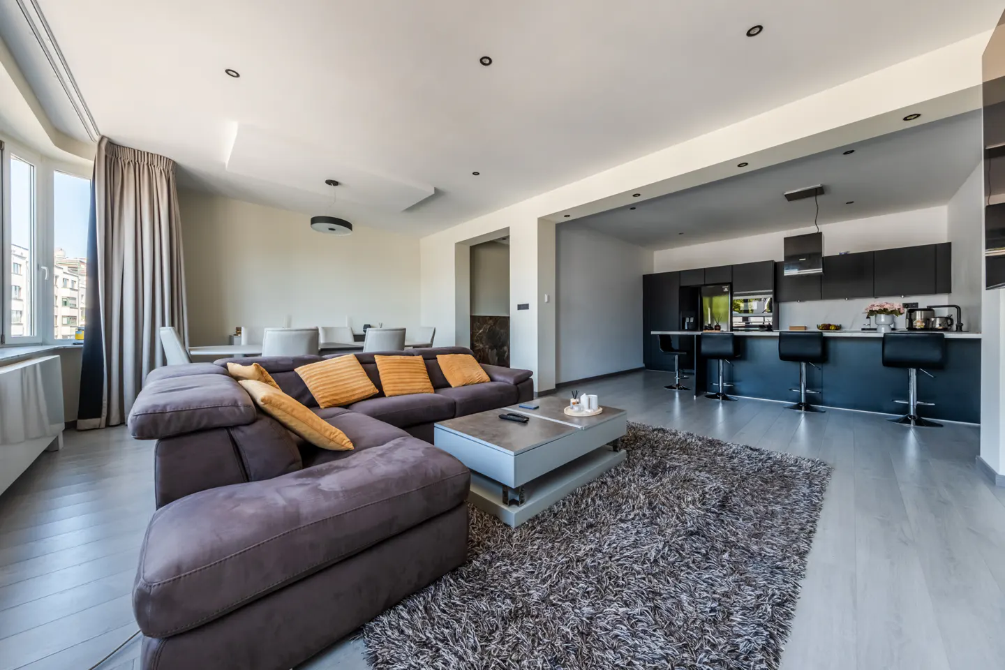 Open-concept living space with a brown sectional sofa, orange pillows, and a gray shag rug. A black kitchen is visible in the background.