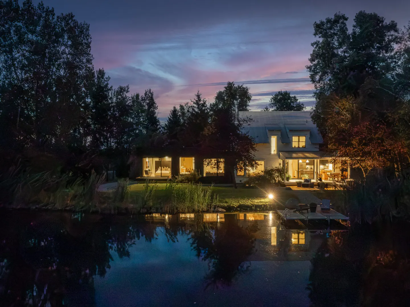 A modern white house with a dock on a lake at dusk, lights reflecting in the water.