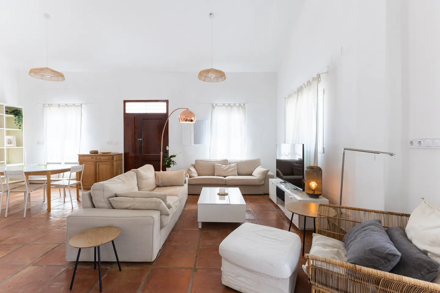 Bright living room with white walls, terracotta tile floor, and beige sofas. Wicker chair, white ottoman, and TV are also visible.