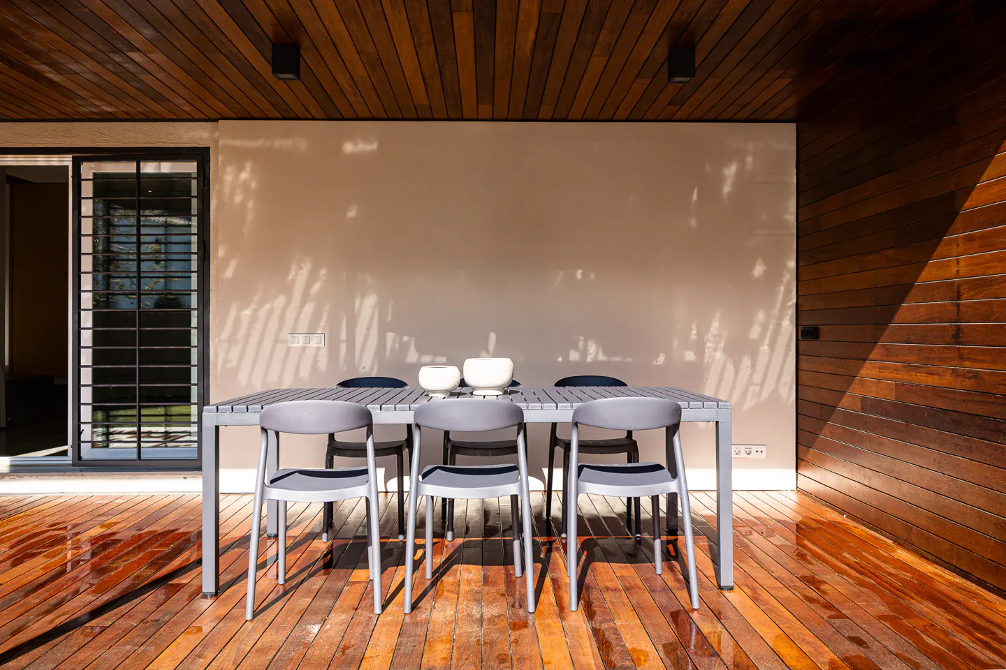 Modern outdoor patio with a grey dining set on a polished wooden deck under a slatted wood ceiling.