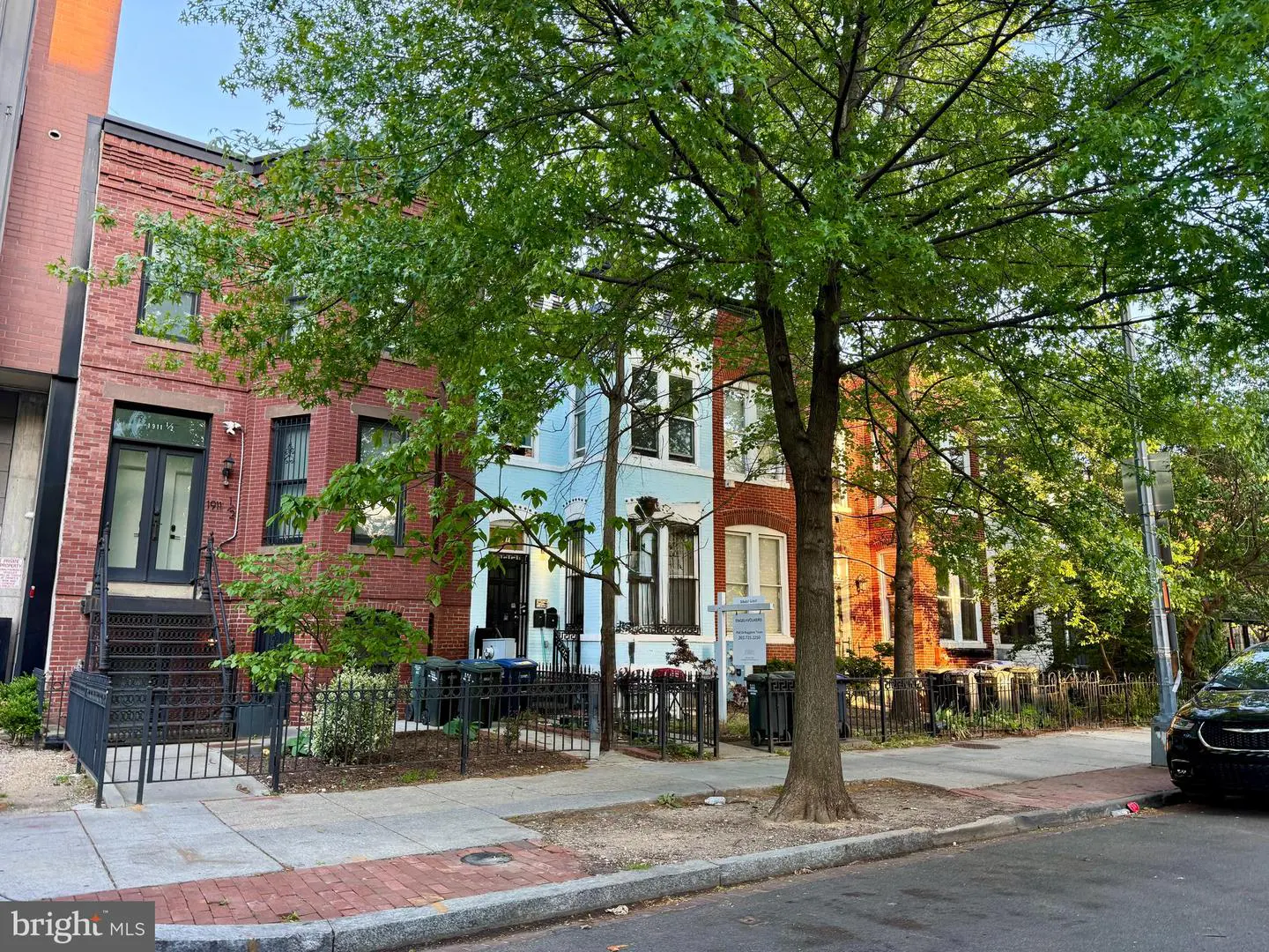 Street view of colorful row houses with brick and blue facades, trees, and black iron fences.