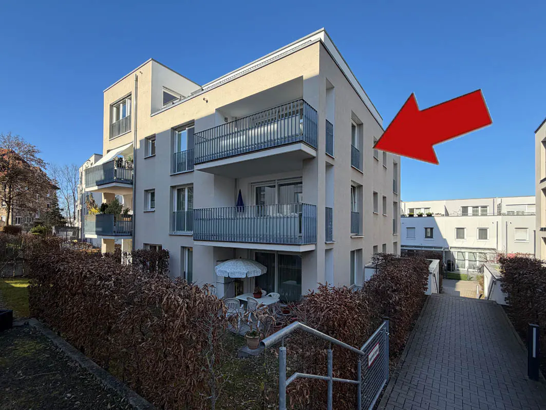 Exterior view of a modern, multi-story apartment building with balconies, a patio set, and a red arrow pointing to the right side of the building.