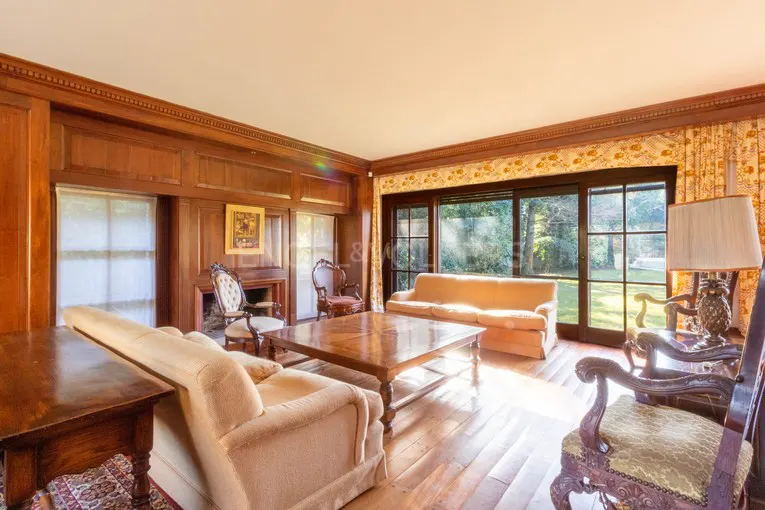A sunlit living room with wood paneling, hardwood floors, and tan sofas. A large window overlooks a green lawn.