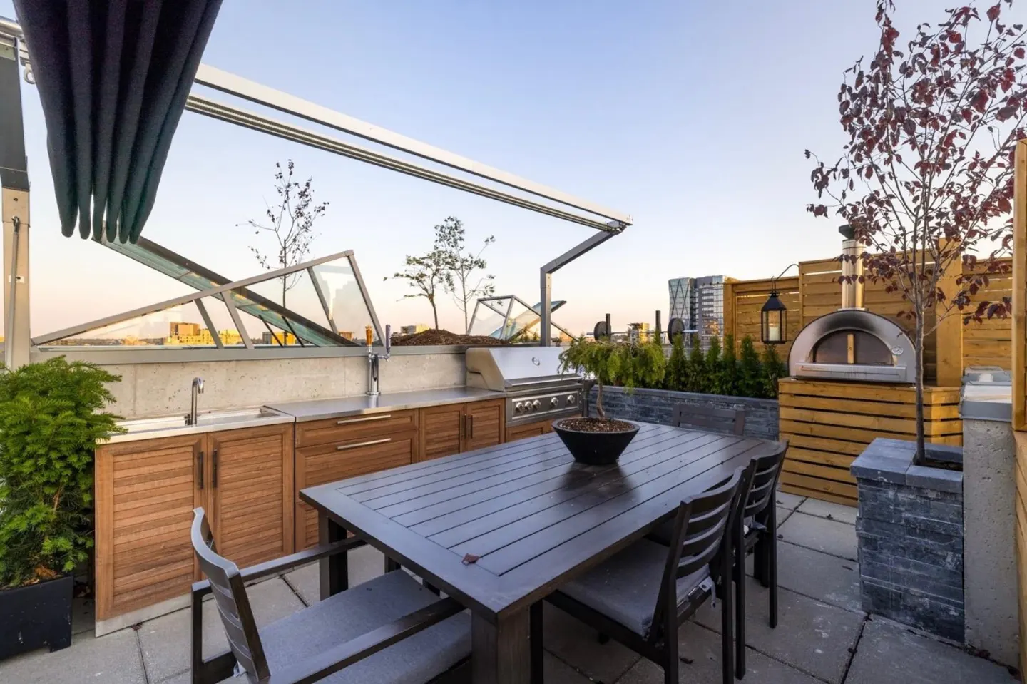 Rooftop patio with outdoor kitchen, dining table, grill, pizza oven, and retractable awning. City skyline in the background.
