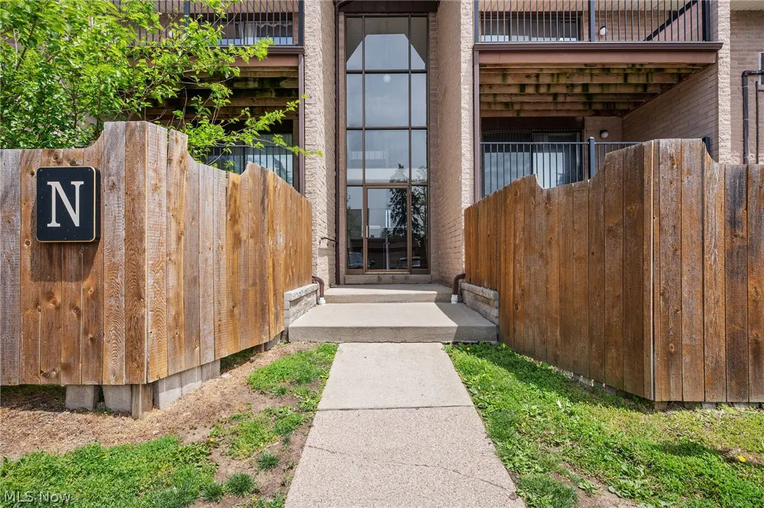 Exterior view of a condo building with a tall glass entrance, flanked by wooden fences and balconies. A concrete walkway leads to the entrance.