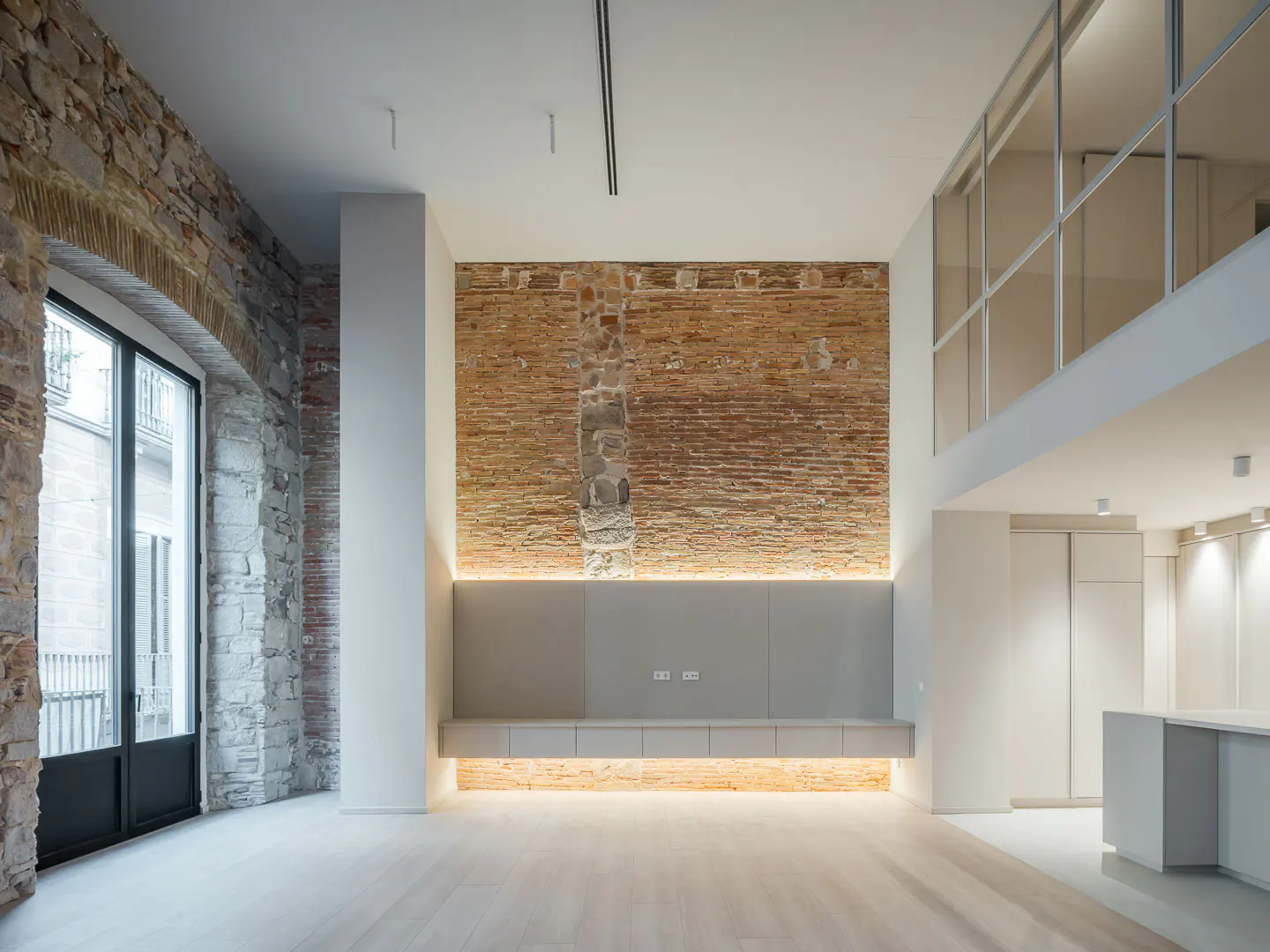 Bright, modern loft with exposed brick wall, light wood floors, and a large arched window. A second-story balcony overlooks the space.