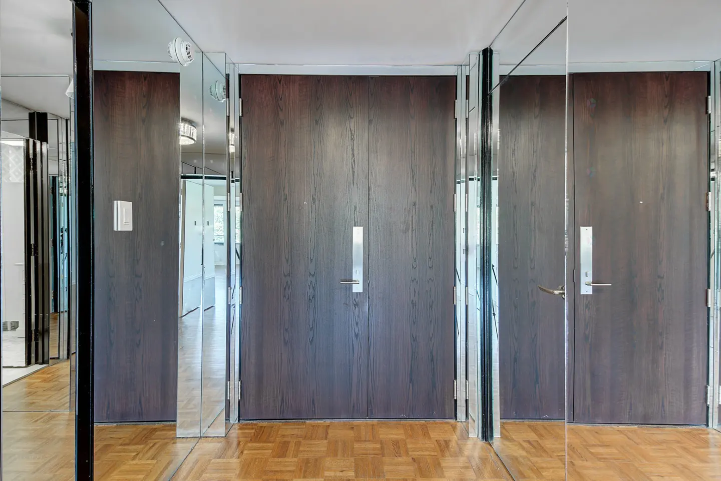 Double dark wood doors with silver handles centered between floor-to-ceiling mirrored walls and parquet wood flooring.