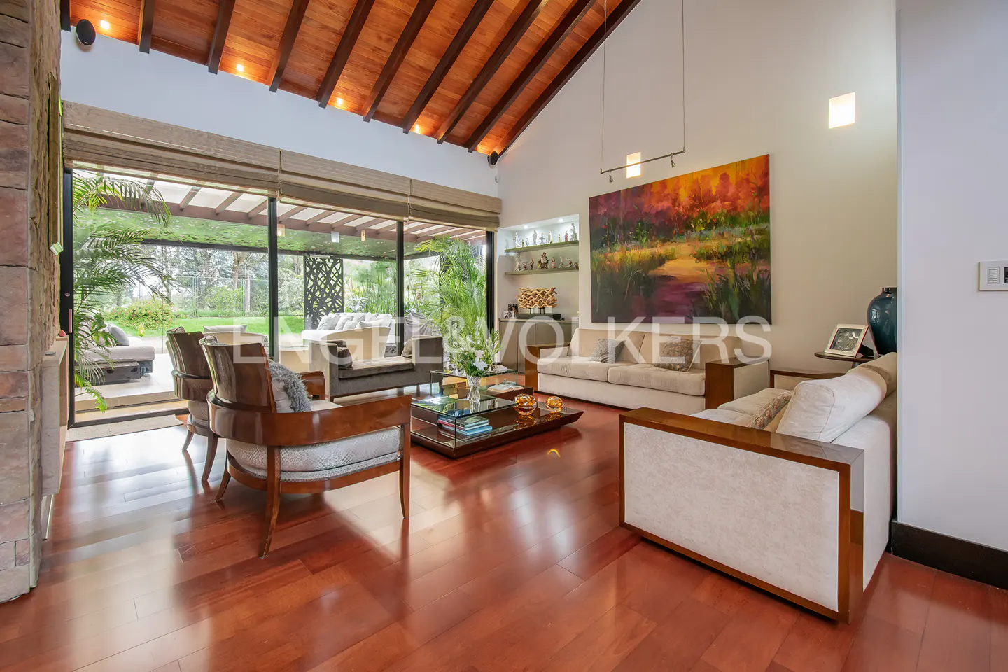 A bright living room with wood floors, a high wood ceiling, and large windows overlooking a green lawn. Two sofas and chairs are arranged around a glass coffee table.