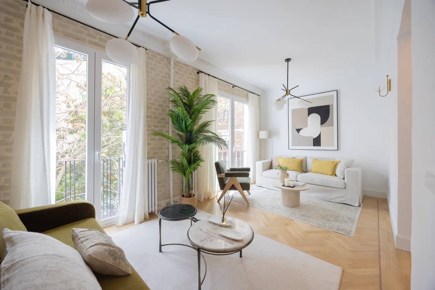 Bright living room with white walls, wood floors, and large windows. Olive sofa, white sofa with yellow pillows, and modern art.