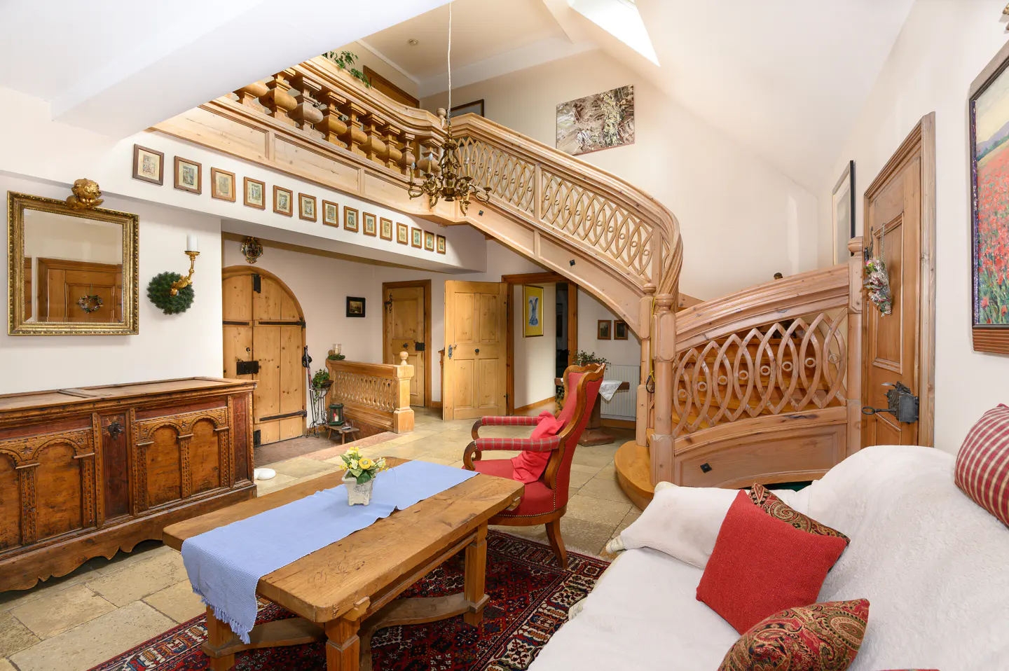 A bright living room with a wooden staircase, table, and sofa. The walls are white, and the floor is tiled.