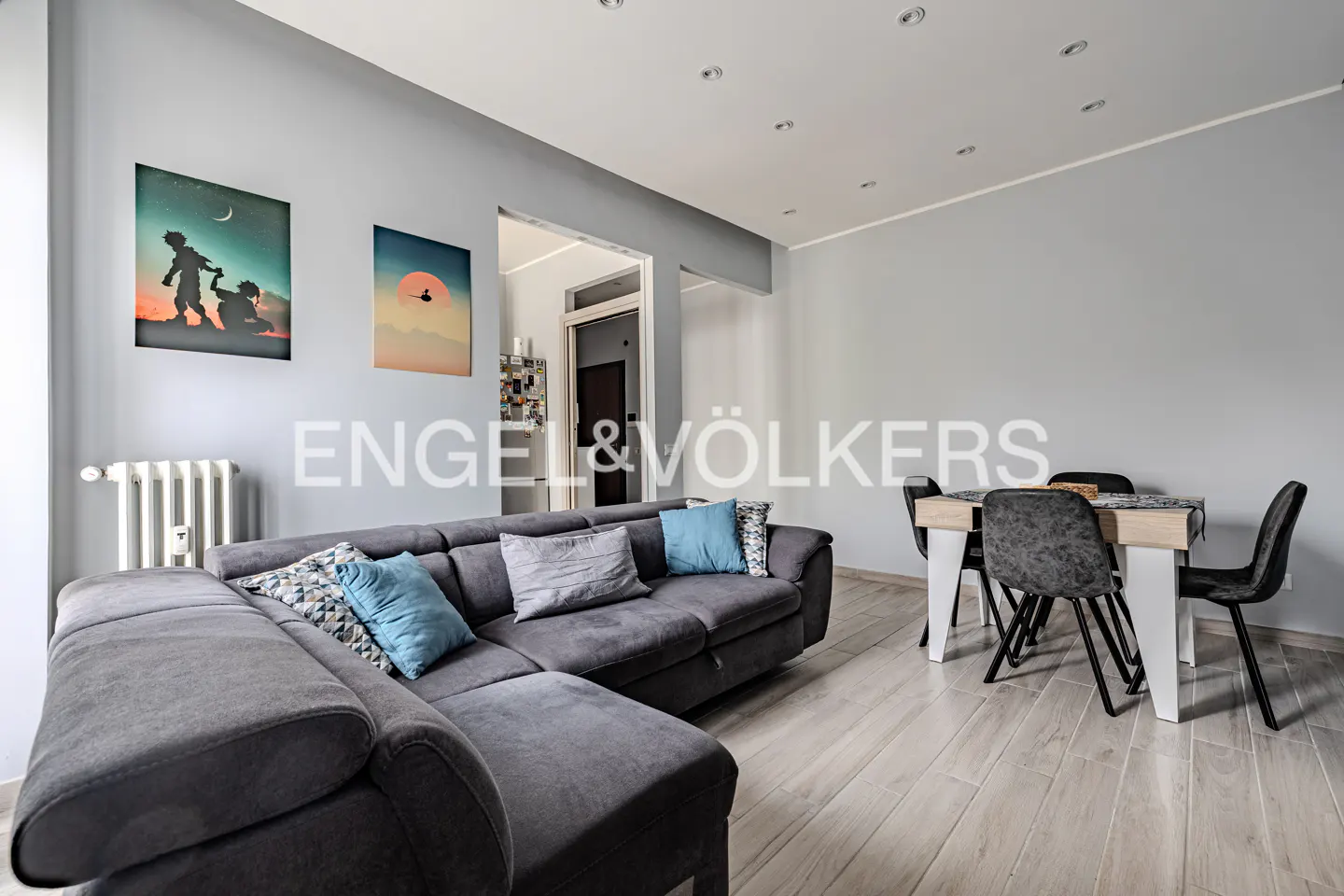 Living room with gray walls, wood floors, and recessed lighting. A gray sectional sofa with blue pillows faces a dining table with four chairs. Two framed prints hang on the wall.