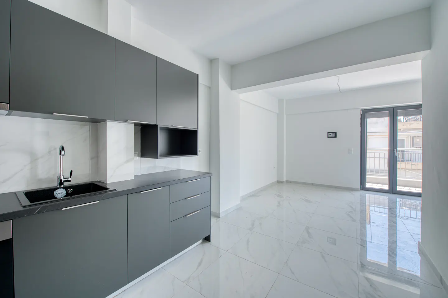 A modern, empty apartment with gray kitchen cabinets, a black sink, and white marble floors. A gray door leads to a balcony.