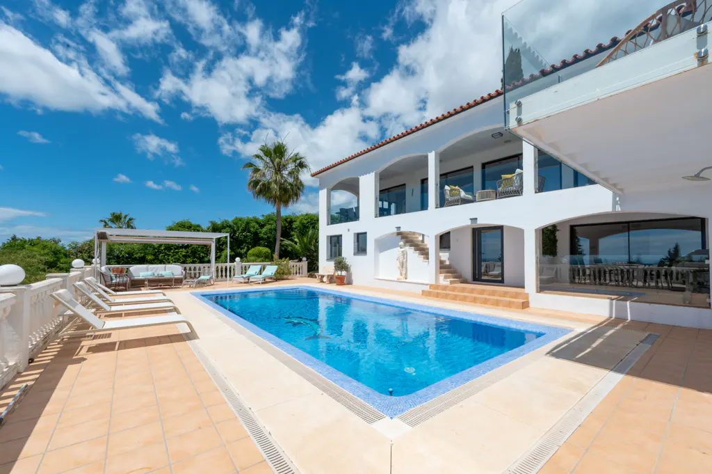 Luxury villa with a blue swimming pool, lounge chairs, and a palm tree under a partly cloudy sky.