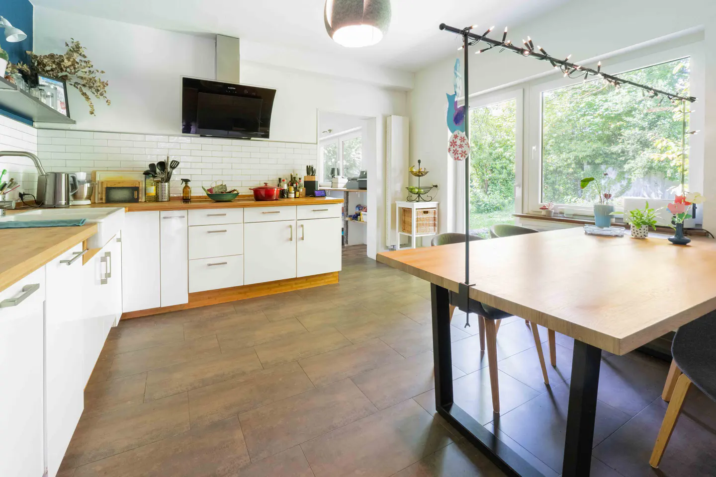 Bright kitchen with white cabinets, wood countertops, and brown tile floor. A large wood table with black legs sits near a window.