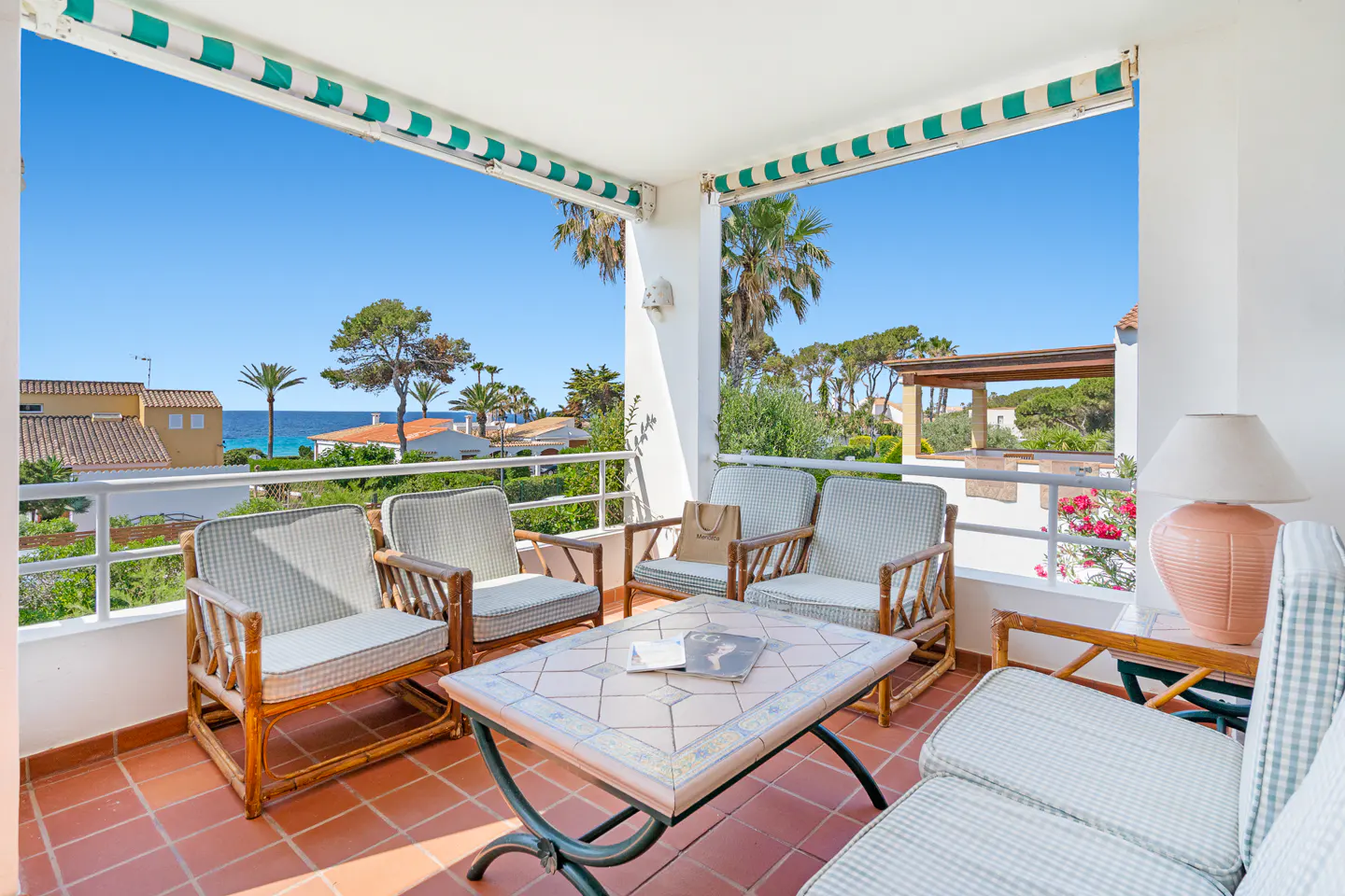 Covered balcony with rattan furniture, tiled table, and ocean view under a green and white striped awning.