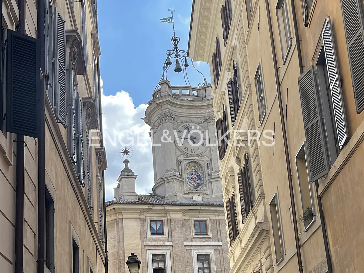 View of a clock tower with bells and a flag, framed by buildings with shutters in Rome, Italy.