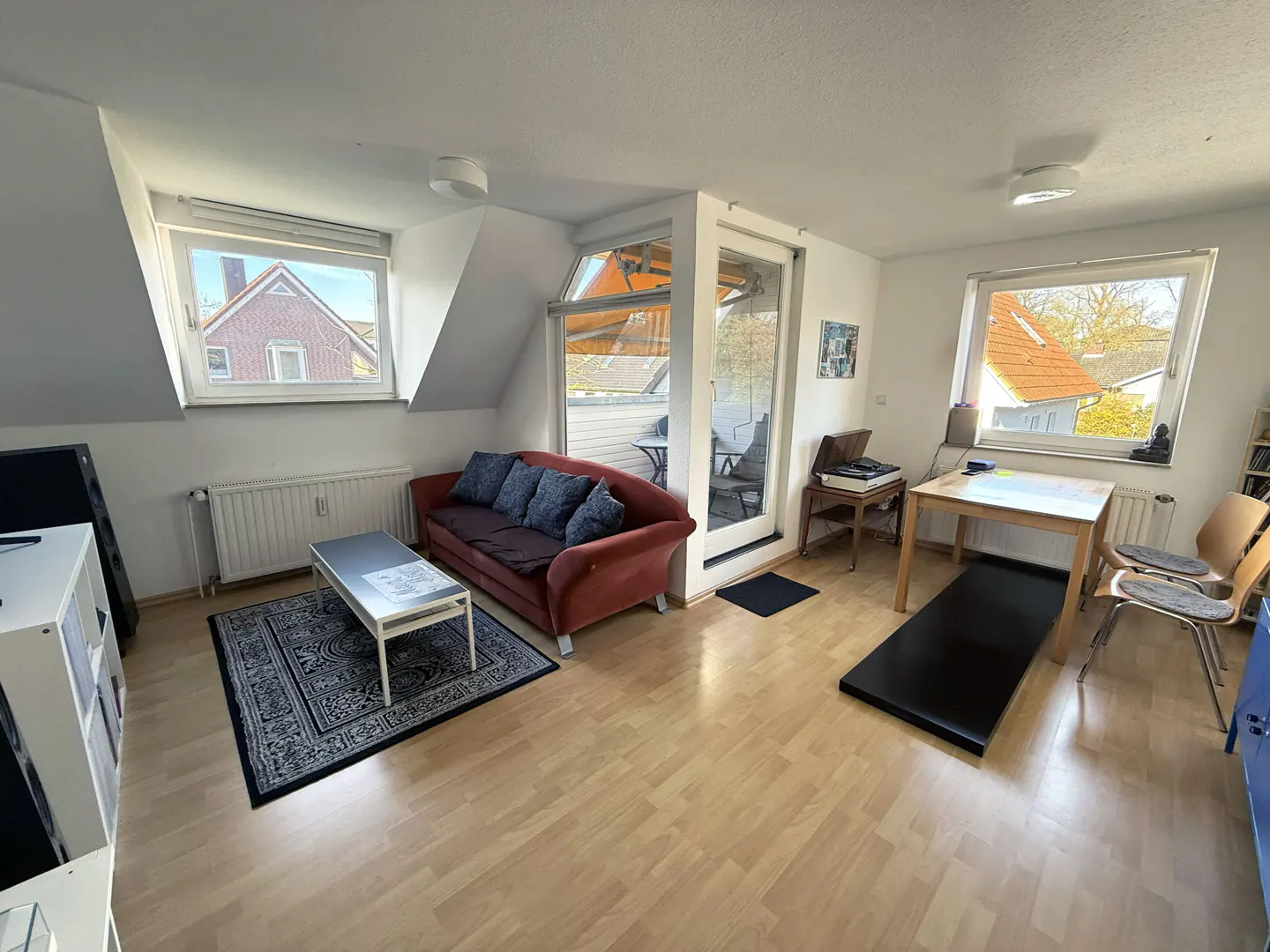 Attic living room with wood floors, a red sofa with blue pillows, a coffee table on a patterned rug, and a table with chairs. Balcony access.