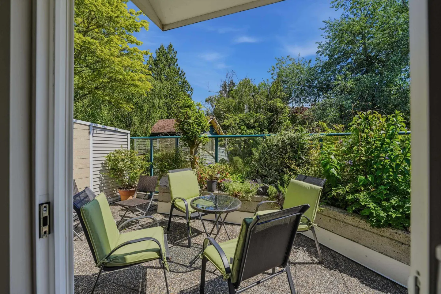 Outdoor patio with green chairs and a glass table, surrounded by lush greenery and a blue sky.