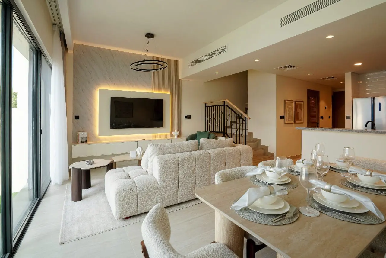 A bright, modern living and dining area with a large white sofa, TV, and a set dining table with plates and glasses.