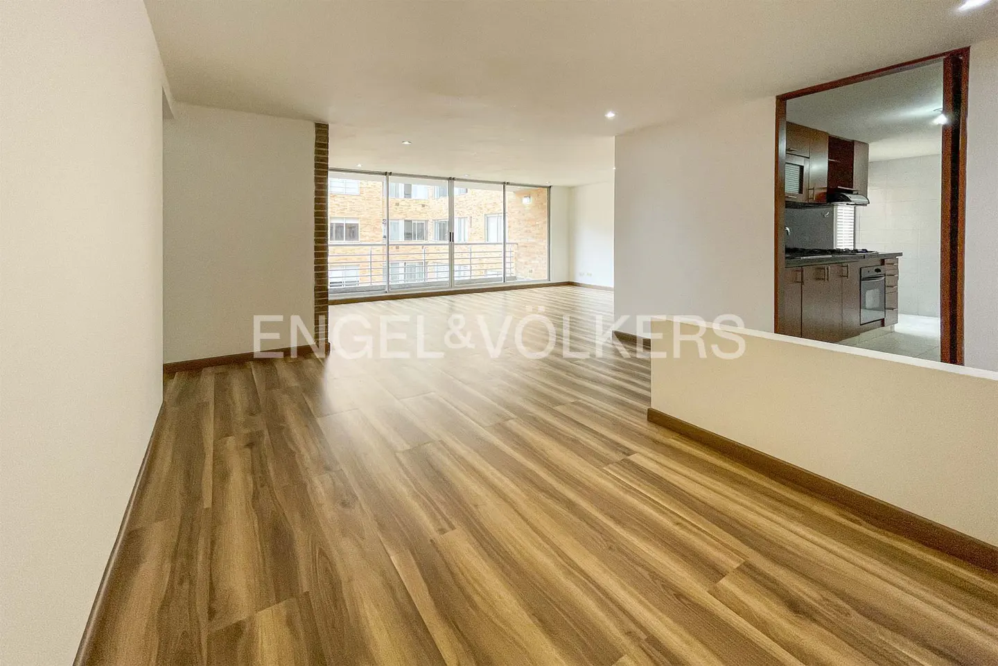 An open-concept living space with wood floors, white walls, and a large window. A kitchen with brown cabinets is visible through a doorway.