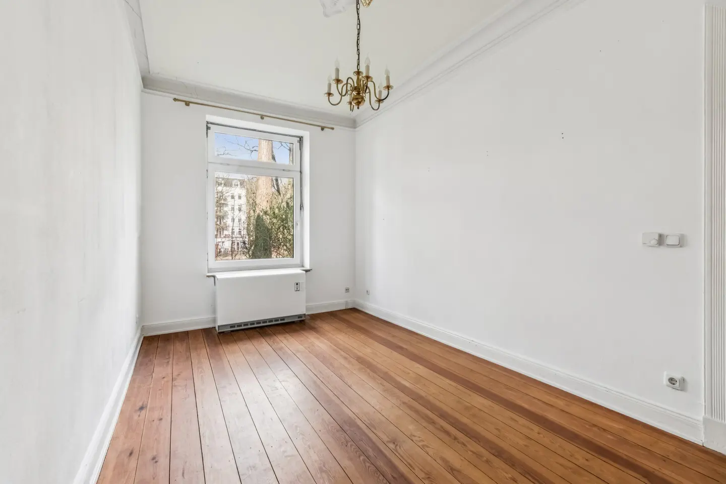 Empty room with hardwood floors, white walls, and a window. A brass chandelier hangs from the ceiling.